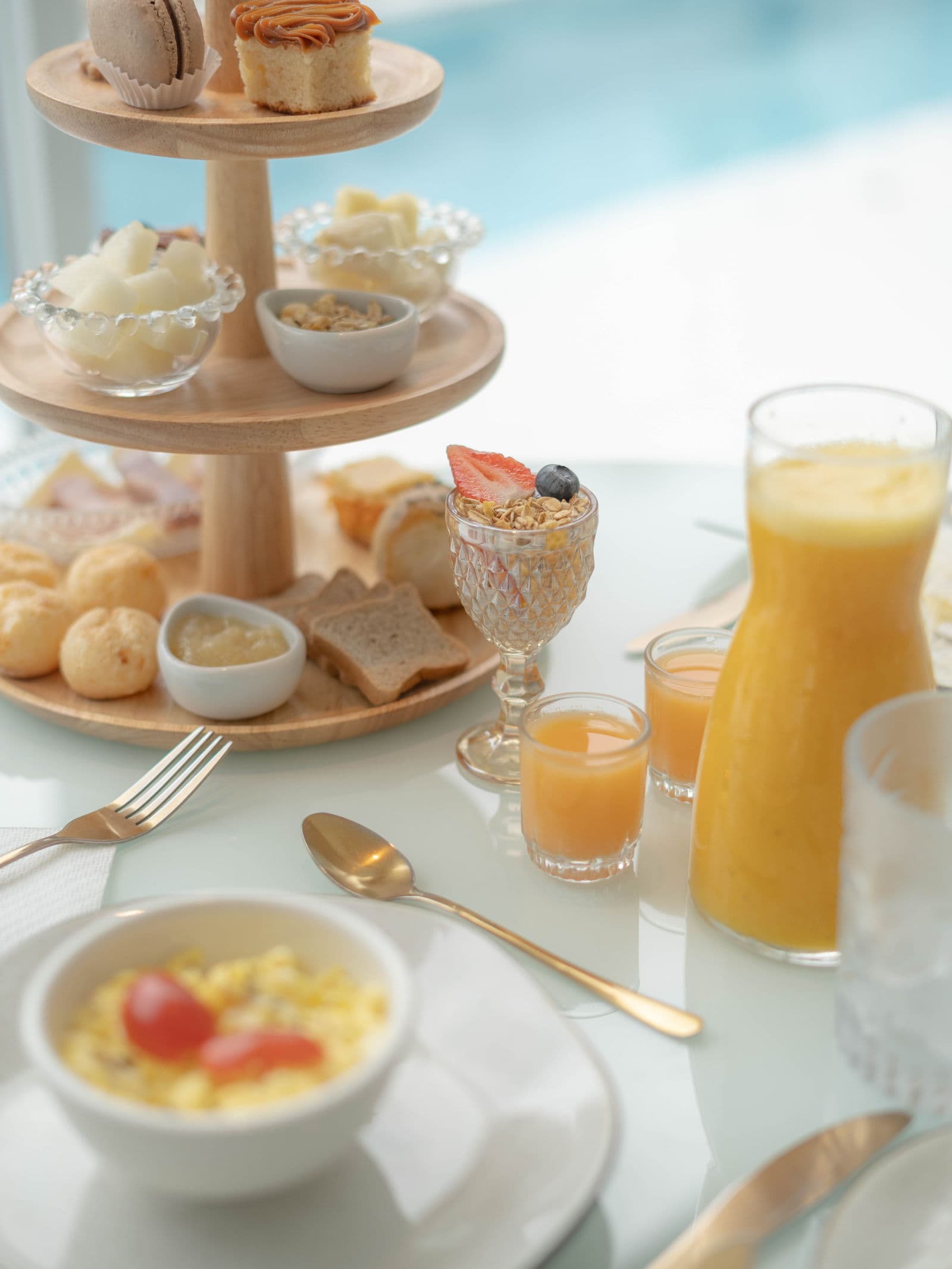 Torre de café da manhã do OLISA com macarons, bolos, pães de queijo e frutas, ao lado de uma jarra de suco de laranja e ovos mexidos