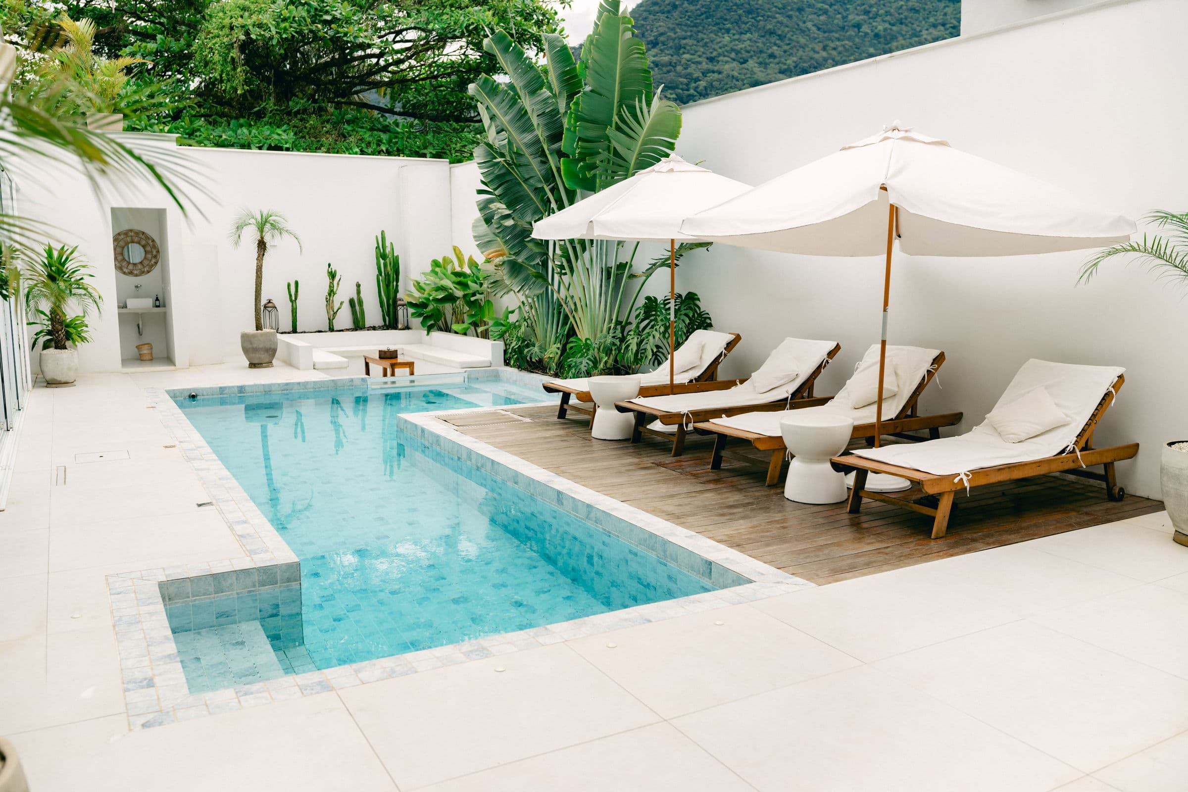 Pátio interno do OLISA com a piscina, o deck de madeira, espreguiçadeiras sob guarda-sóis brancos e a Mata Atlântica atrás do muro caiado