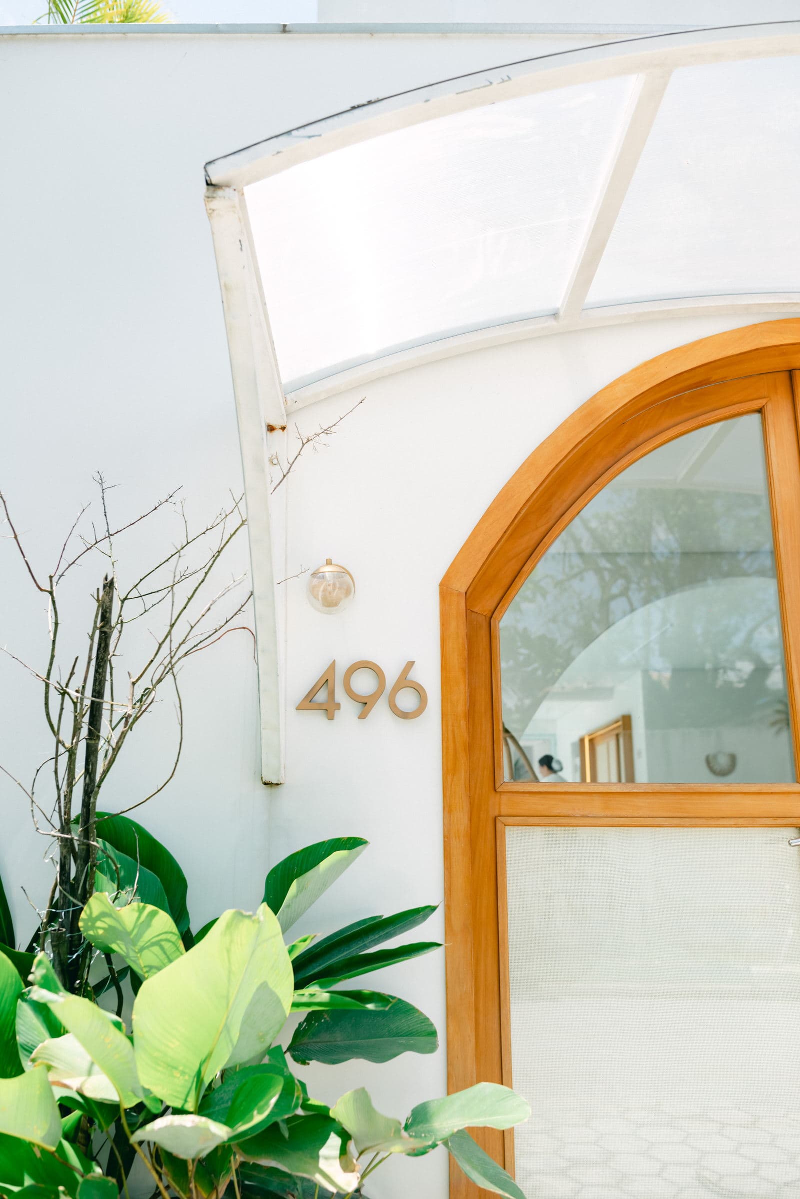 O portal de entrada do OLISA Boutique: porta de madeira maciça em arco e o número 496 em letras douradas sobre o muro branco caiado, entre folhagem tropical
