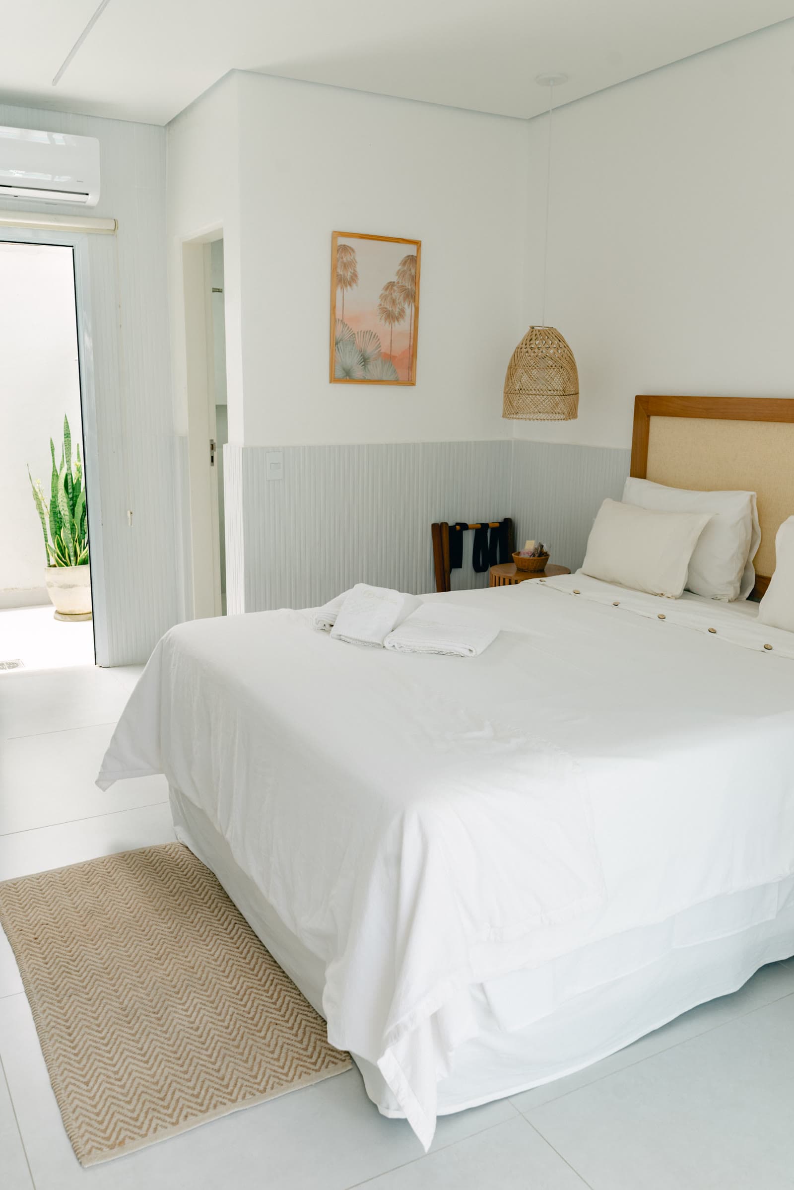 Quarto da Suíte Luxo do OLISA Boutique Hotel: cama de casal com roupa de cama branca e cabeceira de madeira e palha, tapete de fibra natural e a porta aberta para o pátio